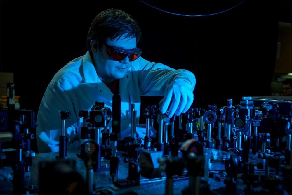 穿着实验室工作服的坦普尔化学教授, 在一个黑暗的实验室里，安全护目镜和手套在许多类似的设备中触摸一个设备.
