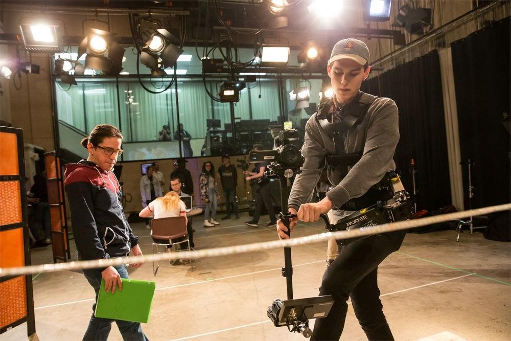 A student in a film studio carefully handles a camera while another student looks on.